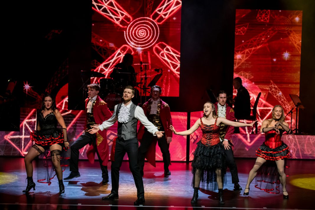 Sieben Darsteller:innen stehen in Rot-Schwarzen kostümen auf der Bühne und Singen