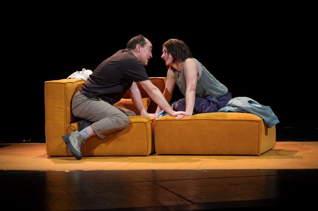 Markus Seuß und Petra-Janina Schultz sitzen zu sich gegenseitig vorgelehnt auf einem Sofa auf der Theaterbühne